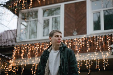 Genç adam ilginç bir bakışla yan tarafa bakar, Yeni yıl çelenkleri ahşap bir eve tutunur, Noel şehrinde dolaşır.