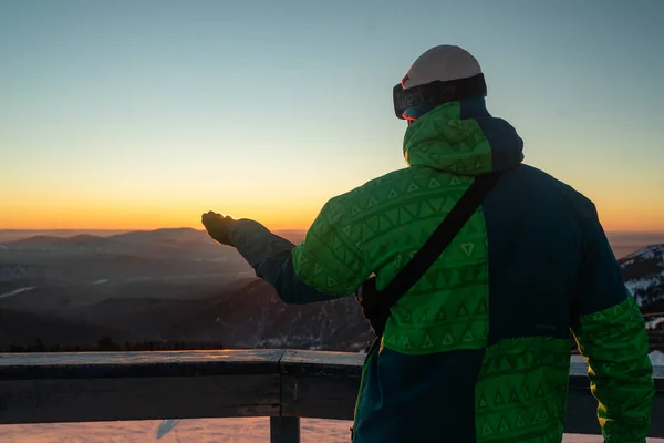 Kayak merkezindeki bir turist, karlı dağların tepesinde durur, elini günbatımına doğru uzatır, karlı dağ zirvelerinin manzaralı doğasının tadını çıkarır.