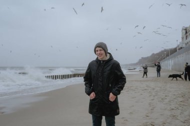 a guy in a winter hat and a warm jacket squints against the strong wind, looks into the camera, portrait of a teenager at the sea, hides his hands in warm pockets