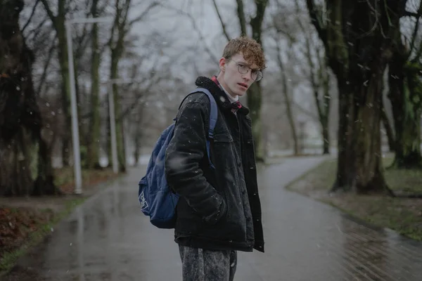 Islak kar, yağmurlu kış, bulutlu hava, kıvırcık saçlı ve görüş için yuvarlak gözlüklü şık genç bir adam parkın ortasında duruyor, sırtında bir sırt çantası ve siyah sıcak bir kot pantolonla kameraya bakıyor.