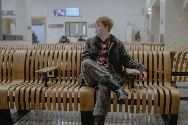 Bir tren istasyonunda, bir havaalanında halka açık bir yerde maske takan genç bir adam seyahat etmek için bekliyor.