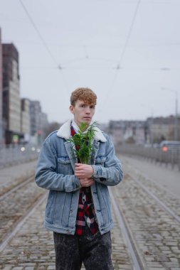 Kıvırcık saçlı bir adam elinde bir saksı tutuyor, göğsüne bastırıyor, hüzünlü bir bakışla, kırgınlıkla izliyor, soğuk bir sonbahar akşamında mavi kot ceket giyiyor.