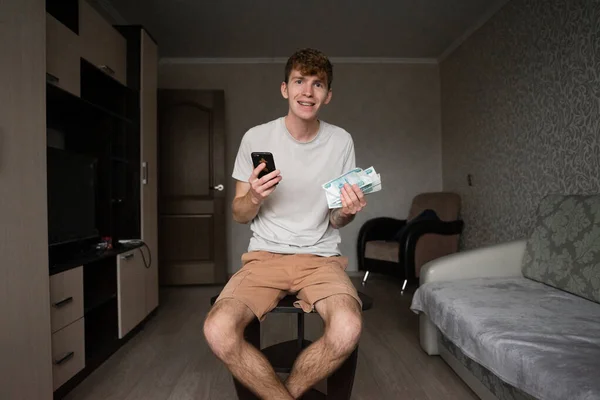 Gri tişörtlü genç bir adam, yüzünde olumlu bir kayıp ile odanın ortasında oturuyor, kameraya bakıyor, elinde bir cep telefonu ve kağıt para tutuyor, internetten kazandığı parayla seviniyor.