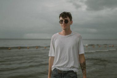 Güneş gözlüklü genç bir adam denizin arka planında duruyor.