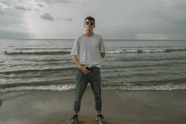 Beyaz tişörtlü ve yuvarlak güneş gözlüklü genç bir adam deniz kıyısında duruyor.
