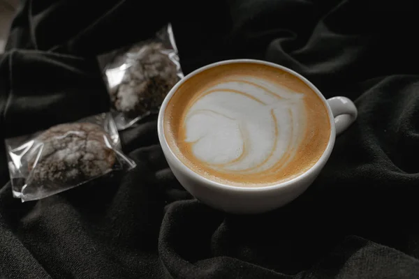 Süt köpüklü bir kahve içeceği, bir fincan kapuçino siyah bir bez üzerinde duruyor, çikolatalı bisküviler yakınlarda yatıyor.