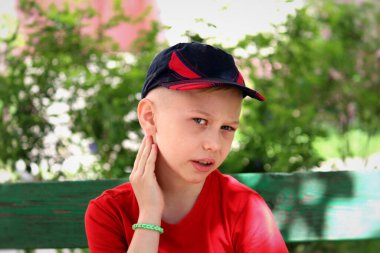 Çocuk bankta caddede oturuyor. Isırığın olduğu yerde elini boynundan tutuyor. Kameraya bakıyor. Bulanık arkaplan.