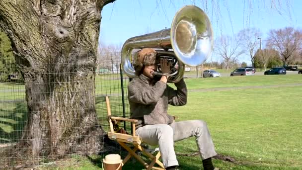 年轻的艺术家戴着棕色的帽子 坐在公园的椅子上 在大号上演奏着缓慢的旋律 — 图库视频影像