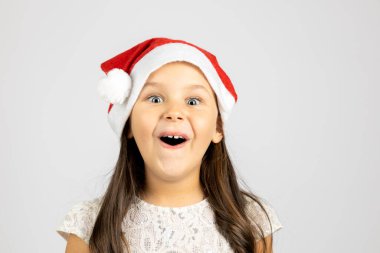 Kırmızı Noel Baba şapkalı, beyaz arka planda izole edilmiş neşeli, komik, ağzı açık, uzun saçlı bir kızın yakın plan portresi.