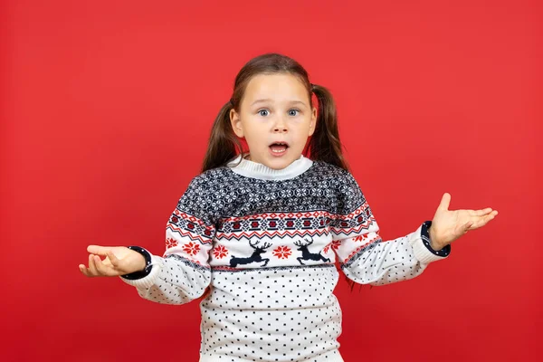 Retrato de chica sorprendida y confundida en suéter blanco de Navidad con brazos extendidos de renos, aislado sobre fondo rojo — Foto de Stock