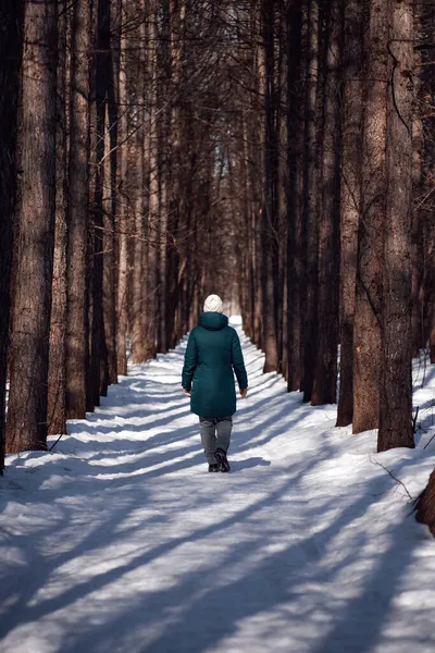 Junge Frau läuft die Straße entlang. Winterliche Straße im schneebedeckten Wald, junge Frau in grüner Daunenjacke spaziert an einem schönen frostigen, sonnigen Tag — Stockfoto