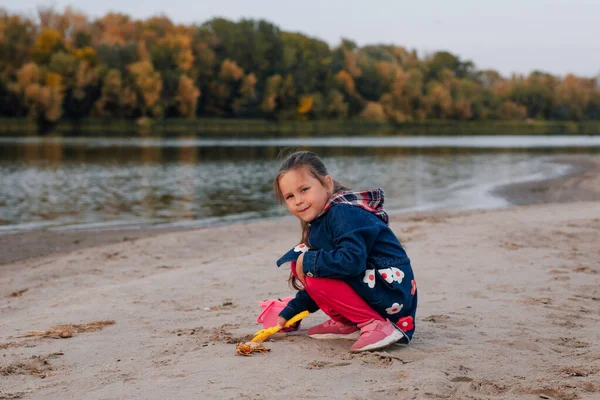 Side view a gyermek játszik a folyóparton ősszel. Lány guggolt le a víz szélén, és felszedett egy maroknyi homokot, hogy hozzáadja a halom előtte, háttér erdő változatos lombozat — Stock Fotó