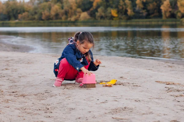 Il bambino sta giocando sulla spiaggia. Bambina in giacca blu e pantaloni rosa sta facendo torte di sabbia sulla riva del fiume nella calda giornata autunnale sullo sfondo della foresta con fogliame colorato — Foto Stock