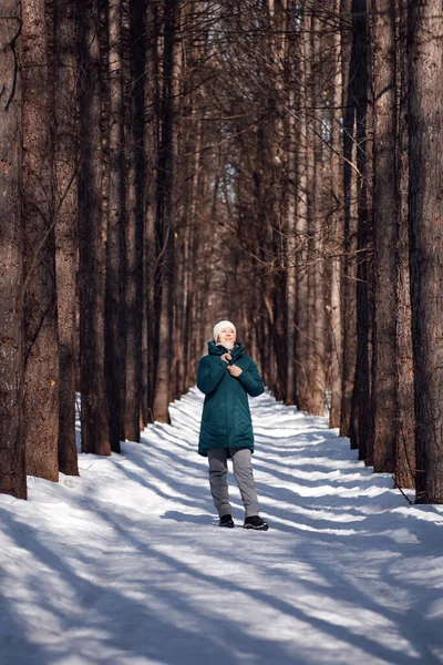 Junge Frau geht durch verschneiten Wald. Junge Frau in grüner Daunenjacke und weißem Hut genießt Spaziergang entlang des Winterweges zwischen hohen Bäumen — Stockfoto