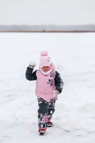 En liten flicka på promenad. Baby i rosa varm kostym spelar snöbollar på snöig väg på bakgrunden av skogen — Stockfoto
