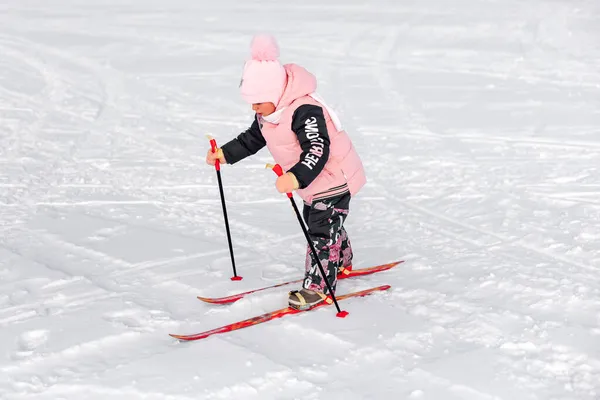 Маленька дівчинка катається на лижах. Дитина в рожевому теплому костюмі вчиться кататися на лижах на засніженій дорозі, зимовий пейзаж, сніговий фон — стокове фото