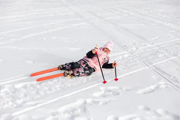 A criança está deitada na neve. Menina feliz em terno rosa está aprendendo a esquiar e caiu na estrada nevada, fundo nevado — Fotografia de Stock