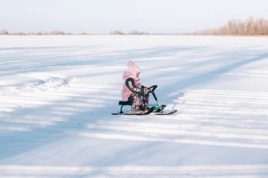 Child rides snowmobile. Little girl in pink warm jacket enjoys walk in nature and sledding on frozen river on sunny winter day clipart