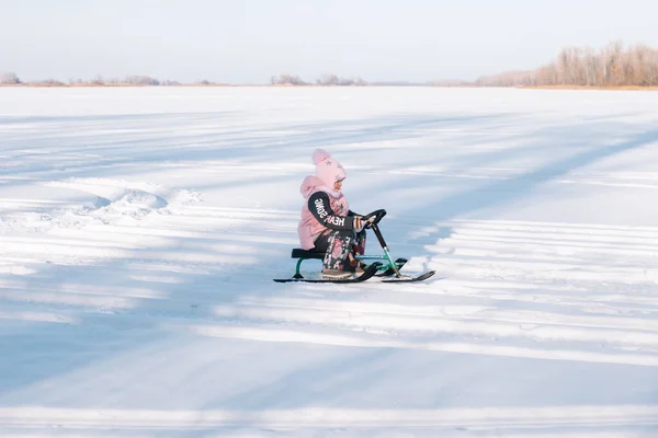 Criança monta snowmobile. Menina no casaco quente rosa gosta de andar na natureza e andar de trenó no rio congelado no dia ensolarado de inverno — Fotografia de Stock