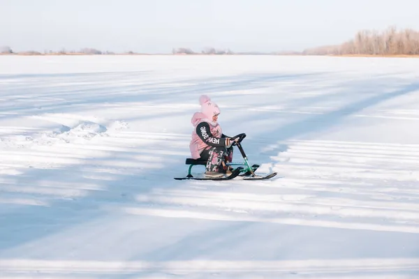 Criança monta snowmobile. Menina no casaco quente rosa gosta de andar na natureza e andar de trenó no rio congelado no dia ensolarado de inverno — Fotografia de Stock