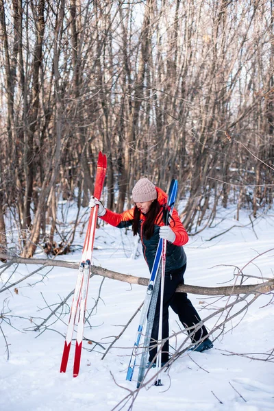 A jovem caminha pela floresta. Morena no traje de inverno caminha ao longo da estrada nevada e detém pólos de esqui e esquis em suas mãos — Fotografia de Stock