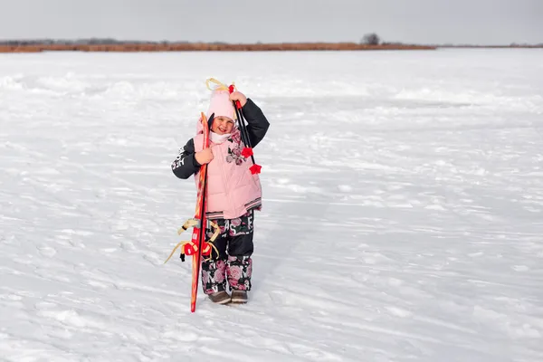 A menina está a esquiar. Criança no fundo da floresta de inverno está de pé na estrada coberta de neve, segurando crianças esquis em suas mãos e olhando para a câmera — Fotografia de Stock
