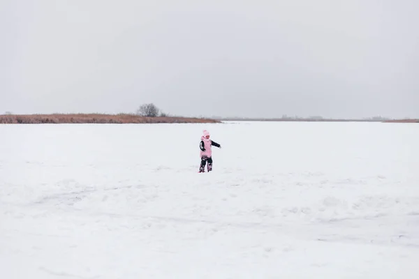 Kind vor dem Hintergrund der Winterlandschaft. Kleines Mädchen in Anzug und Hut mit Bommel steht auf schneebedecktem Eis mitten im See — Stockfoto