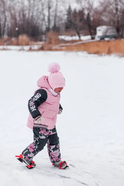 Ragazzina a piedi. Ragazzina felice in abito caldo rosa cammina su racchette da neve sulla strada innevata vicino alla riva del fiume ghiacciato, vista laterale — Foto Stock