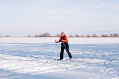 Esmer, donmuş nehirde kayak yapıyor. Kışlık takım elbiseli genç kadın ormanın arka planında spor yapıyor, kayak yapmayı öğreniyor, yandan bakmayı.