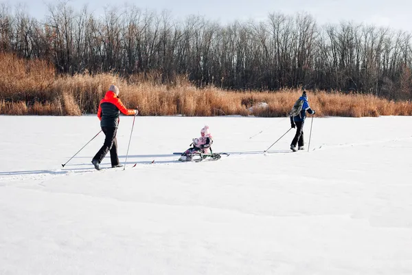Família em passeio de inverno em dia ensolarado. Jovem mulher, homem e criança estão esquiando no rio congelado no fundo da floresta, o homem está montando criança no trenó das crianças na neve — Fotografia de Stock