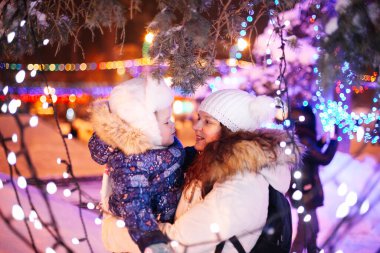 Genç bir kadın Noel ağacının altında Noel pazarıyla süslenmiş bir meydanda küçük bir kızı kollarında tutuyor.