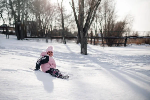 Barnet leker i parken på vintern. Liten flicka rider snörutschbana och njuter av varm solig dag på vintern semester — Stockfoto