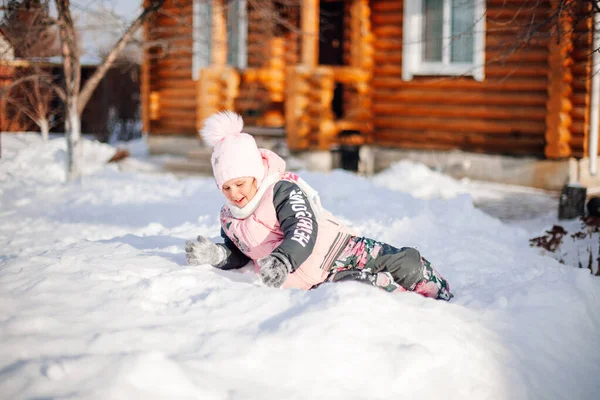 Маленька дівчинка лежить на снігоході. Дитина розважається в сонячний зимовий день на задньому дворі на фоні дерев'яного будинку, грає в сніжки, стрибає в сніг — стокове фото