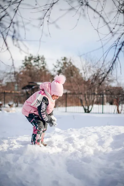 Kind spielt in der Natur. Kleines Mädchen im warmen Anzug spielt im Hinterhof und genießt sonnigen Tag im Winter, sie steht auf Schneewehen und spielt Schneebälle, Seitenansicht — Stockfoto