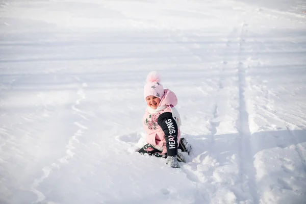 La petite fille est assise dans la neige. Enfant en veste d'hiver rose et chapeau avec pompon s'amuse sur la promenade dans la nature et bénéficie d'un temps ensoleillé et joue avec la neige le matin d'hiver givré — Photo