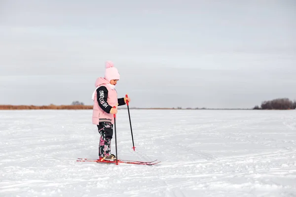 A menina está a aprender a esquiar. Criança de terno quente rosa esquiando na neve no dia gelado de inverno, vista lateral, fundo de neve — Fotografia de Stock
