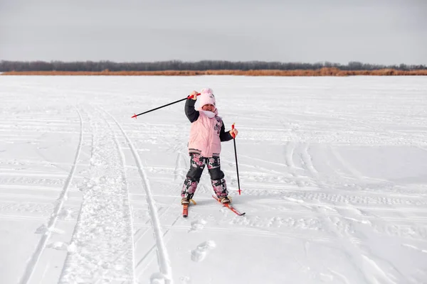 Criança está esquiando na neve. Menina feliz em traje quente aprende a esquiar na margem do rio congelado e acena sua mão, paisagem de inverno, fundo nevado — Fotografia de Stock