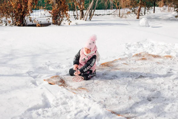 La fille est assise sur le tapis en hiver. Enfant apprend à nettoyer le tapis par la méthode traditionnelle à l'aide de neige fraîche, elle utilise un balai pour balayer la neige — Photo
