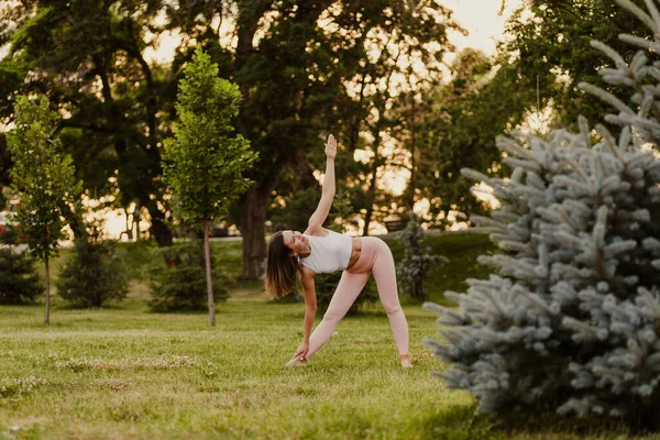 Attractive, elegant woman do yoga and stands in Trikonasana pose while maintaining balance on green grass of lawn at sunset, concept of inner harmony — Stock Photo, Image