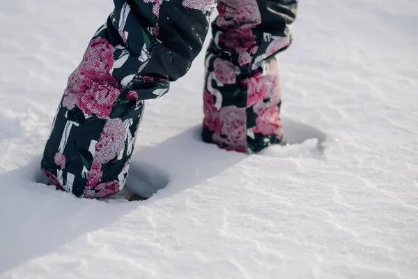 L'enfant marche à travers la neige. Vue rapprochée des jambes des enfants en pantalon chaud avec l'image de fleurs marchant le long d'une route enneigée après de fortes chutes de neige par une matinée ensoleillée et givrée — Photo