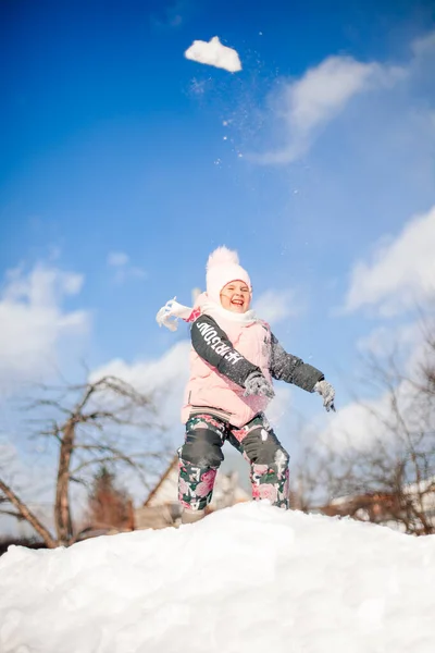 Kind wirft Schneebälle in Winterpark Kleines Mädchen im rosafarbenen Winteranzug spielt und reitet an einem sonnigen Wintermorgen die Schneerutsche vor blauem Himmel hinunter — Stockfoto