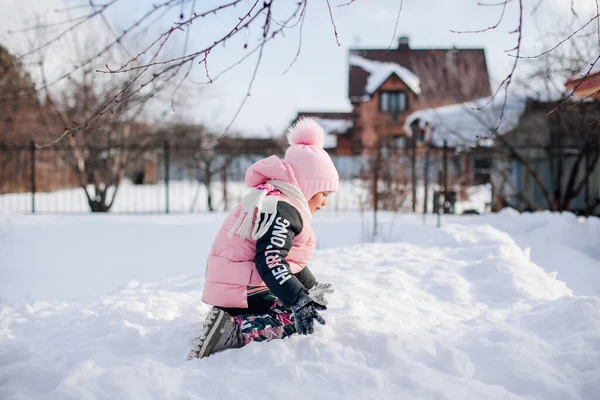La bambina fa pupazzo di neve. Bambino in tuta calda divertirsi nella soleggiata giornata invernale in cortile sullo sfondo della casa di legno, giocando palle di neve, vista laterale, paesaggio invernale — Foto Stock