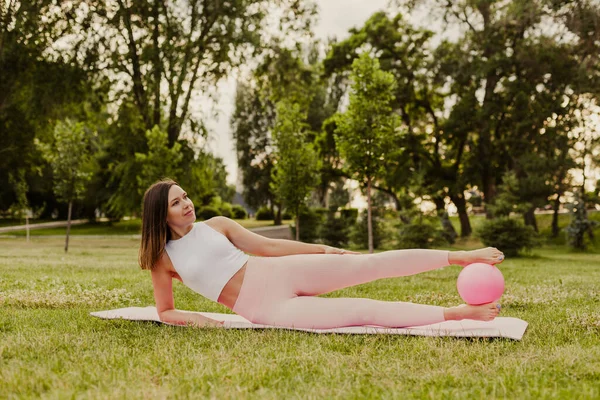Mujer tranquila y sonriente en ropa rosa y una pequeña bola de goma haciendo pilates en alfombra rosa al atardecer en el parque en verano —  Fotos de Stock