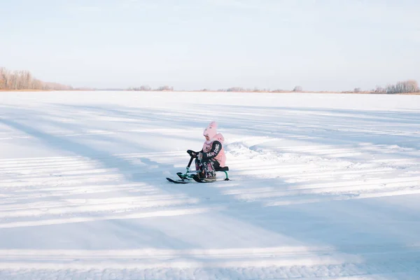 Enfant monte snowcat sur route enneigée. Petite fille en veste chaude rose jouit d'une promenade dans la nature et de la luge sur la rivière gelée par une journée d'hiver ensoleillée, vue latérale — Photo
