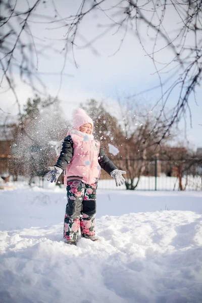 Glückliches Kind auf Winterwanderung. Kleines Mädchen im warmen Anzug spielt im Hinterhof und genießt den sonnigen Tag im Winter, sie steht auf Schneewehen und spielt Schneebälle — Stockfoto