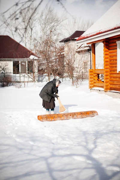 Femme blanche nettoie tapis. Femme âgée en écharpe et bottes en feutre balaie la neige du tapis dans la cour arrière près de la maison en bois, elle est engagée dans le nettoyage de l'environnement — Photo