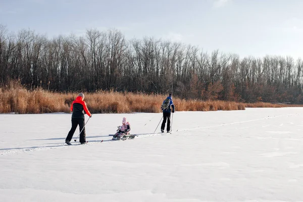 Família em passeio de inverno. Jovem mulher, homem e criança estão esquiando no rio congelado perto da floresta, o homem está montando criança no trenó das crianças na neve — Fotografia de Stock