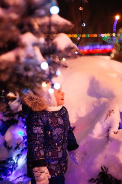  Kışın giyinmiş küçük bir kız Noel ağacının altında kar yığınında duruyor. Çelenklerle süslenmiş, Noel süsleri, kutlama ve eğlence atmosferi.