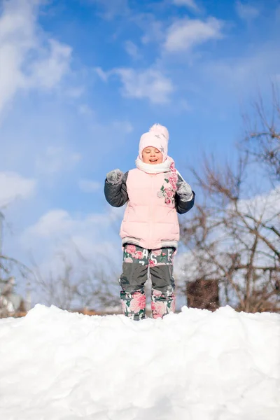 Den lilla flickan leker i naturen. Barn i rosa vinter jacka och hatt med pompom har kul på solig vinterdag, leker och rider ner snörutschbana utan släde och hoppar in i snödrivor — Stockfoto