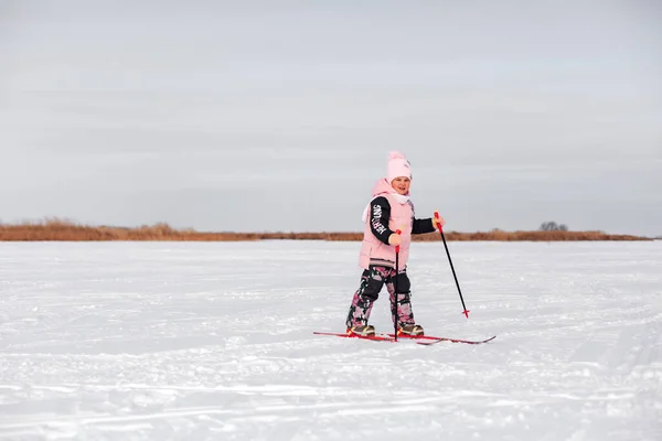 A criança aprende a esquiar. Menina em rosa terno quente esqui na neve no dia de inverno gelado, vista lateral, fundo de neve — Fotografia de Stock